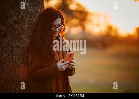 A woman with long, dark hair stands near a tree, using her smartphone. The setting sun bathes the scene in warm, golden light. Stock Photo