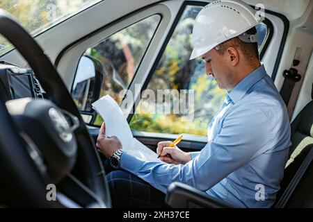A construction worker wearing a hard hat sits in a white van, reviewing documents and writing notes. Stock Photo