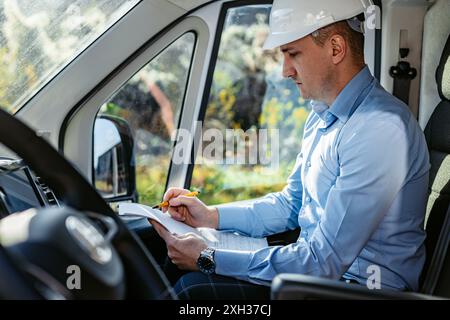 A construction worker wearing a white hard hat sits in the driver's seat of a van, writing in a notebook with a yellow pencil. Stock Photo