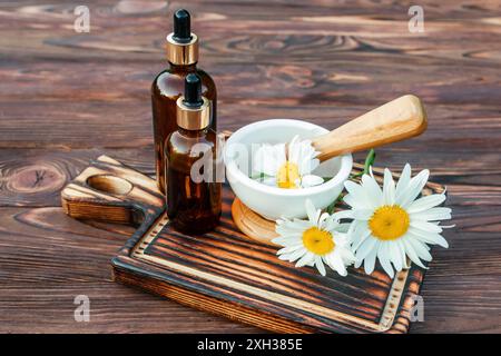 Three brown glass bottles with droppers sit beside a white mortar and pestle with chamomile flowers inside on a burnt wooden cutting board. The cuttin Stock Photo