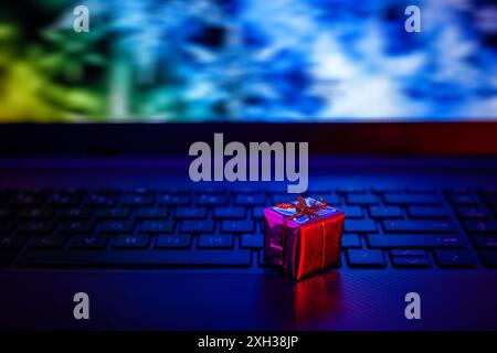 A small, red gift box with a gold bow sits on a black laptop keyboard. The laptop's screen is out of focus, showing blue and green colors. Stock Photo