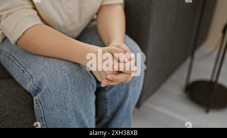 A close-up of a young woman’s folded hands resting on her lap in a casual home setting. Stock Photo