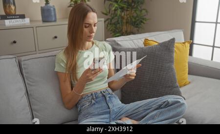 A young, attractive blonde woman sits in a cozy living room with modern decor, holding a smartphone and a piece of paper, looking focused and engaged. Stock Photo