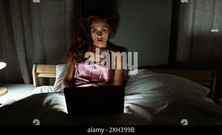 A young caucasian woman works on a laptop in her bedroom at night, creating a scene of focus and solitude. Stock Photo