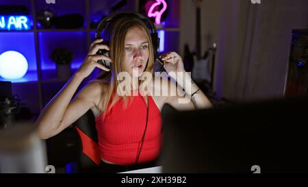 A young woman with headphones in a gaming room at night, focused on a computer screen with neon lights in the background. Stock Photo