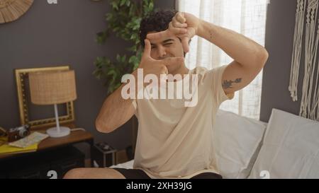 A handsome hispanic man sitting on a bed in a cozy bedroom making a frame with his fingers, creating an inviting and comfortable indoor home setting. Stock Photo