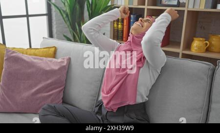A joyful hispanic woman in a hijab sits relaxing in a cozy living room, embodying comfort and happiness at home. Stock Photo