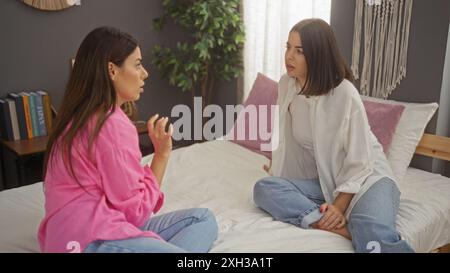 Two women sitting on a bed having a serious conversation in a cozy bedroom, indicating a setting of friendship, intimacy, and trust. Stock Photo