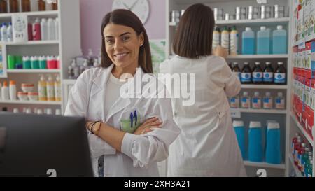 Two smiling hispanic women pharmacists in white coats working together in a well-organized drugstore interior, one facing the camera, and the other ar Stock Photo