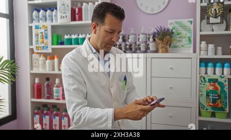 Middle-aged hispanic man in a pharmacy checking his phone while wearing a white lab coat surrounded by shelves of various products Stock Photo