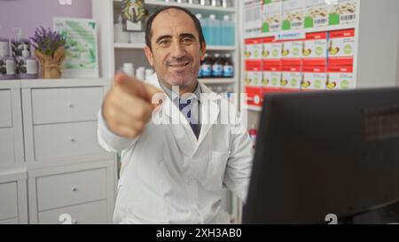 Mature hispanic male pharmacist in a white coat pointing and smiling at a pharmacy counter with shelves stocked with various medications Stock Photo