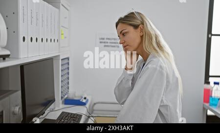 A contemplative young woman in a white lab coat analyzes data on a computer in a bright, modern laboratory setting. Stock Photo