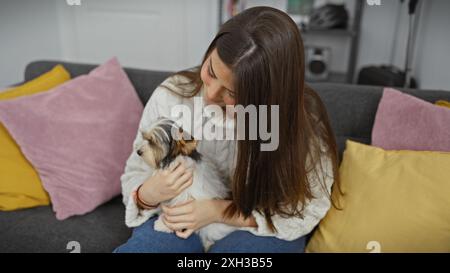 A young woman lovingly holds a small biewer terrier in a cozy living room setting with colorful cushions. Stock Photo