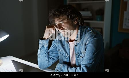 A pensive young man in a denim jacket studying a document at night in a home office setting. Stock Photo