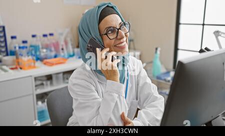 A smiling woman in a hijab and lab coat talking on a phone in a laboratory setting. Stock Photo