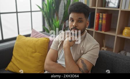 A handsome african man relaxes thoughtfully on a couch in a cozy living room setting with plants and books. Stock Photo