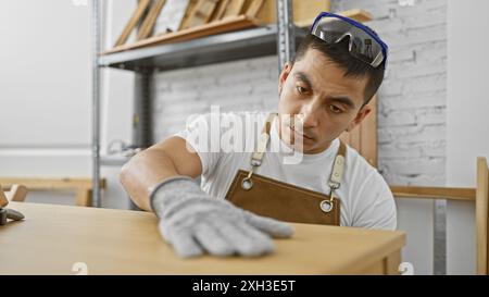 A focused young hispanic man wearing a white shirt and apron examines furniture in a well-lit woodworking workshop. Stock Photo