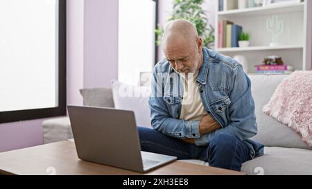 Mature bald man with beard feeling abdominal pain at home, sitting by laptop in living room. Stock Photo