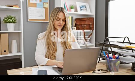A focused blonde woman works on a laptop in a modern office, surrounded by organized shelves and office supplies. Stock Photo