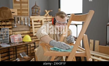 A bearded young man wearing safety glasses and earmuffs sands a wooden chair in a well-equipped carpentry workshop. Stock Photo