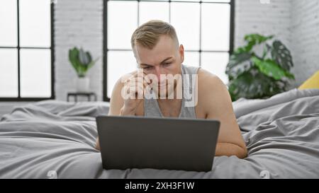 A focused young man with a beard works on his laptop in the comfort of his modern bedroom. Stock Photo