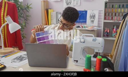 A young man compares fabric colors during a video call in his tailor shop, surrounded by design tools. Stock Photo