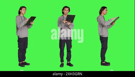 Asian adult reading information on files, thinking about annotations on clipboard papers or taking notes for finalization. Young guy writing on documents, standing against greenscreen. Stock Photo