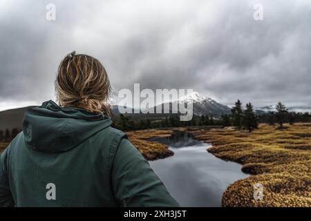 Rear view of a woman in a green jacket by a calm mountain river gazes at a snow covered mountain peak in Nordic Lapland, storm clouds looming overhead Stock Photo
