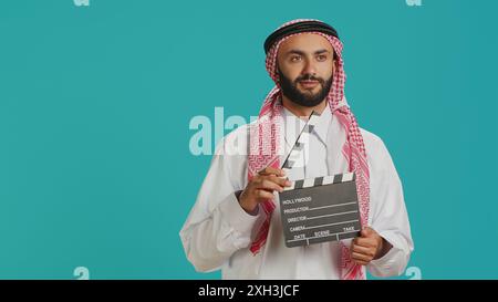 Middle eastern man holds clapperboard used for action takes on movie production, male producer using cinematography film slate. Muslim filmmaker wearing traditional arabic clothing. Stock Photo