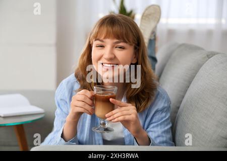 Happy young woman with glass of tasty iced latte lying on sofa at home Stock Photo