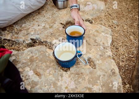 A close-up of two enamel mugs placed on a rock outdoors. One is empty, and the other contains a hot beverage. A hand reaching for the mug suggests a c Stock Photo