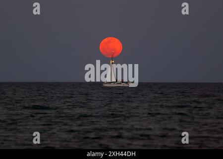 A sailboat crosses in front of a full blood moon as it rises over the Atlantic ocean Stock Photo