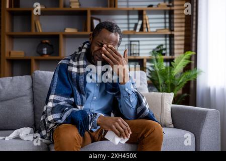 Man feeling unwell sitting on sofa covered with blanket holding tissues. Displaying symptoms of cold or flu, gripping face in discomfort. Home environment with bookshelves and plants in background Stock Photo