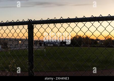 Sunset hues backlighting a chain-link fence - golden hour sky gradient transitioning from warm orange to cool blue - silhouette of urban skyline in th Stock Photo