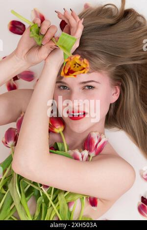 Lovely blonde lady lying with bouquet of spring tulips for Valentine's Day, covering her chest. Gorgeous young blonde is looking at camera with loving Stock Photo