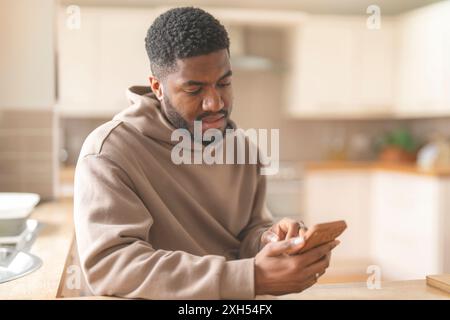 African american man in brown hoodie  using phone, studying, learning, shopping, working online, talking while sitting on a couch at home Stock Photo