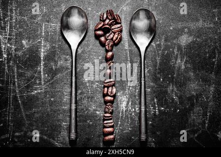 Two metal spoons lie parallel on a textured, dark surface. A vertical line of coffee beans is arranged between them forming an abstract spoon shape. Stock Photo