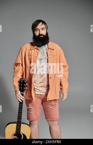 A man stands against a grey background, holding a guitar. He is dressed in a coral-colored jacket, shorts, and a tie-dye shirt. Stock Photo