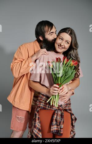 A man and woman in sophisticated attire pose together in a studio setting. The man kisses the woman on the cheek, and she holds a bouquet of red tulip Stock Photo
