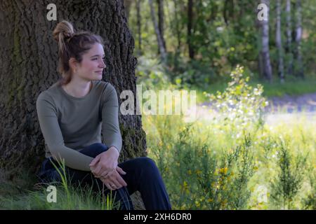 A young woman sits under a tree, finding peace and calm in the serene natural beauty on a sunny day Stock Photo