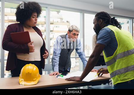 Adult African Man with dreadlocks and reflective clothing is standing explaining to his boss plans of work with plan on table. It is meeting with thre Stock Photo