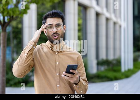 Surprised man holding smartphone outdoors near office building. Young businessman with glasses in casual clothing making phone call, expressing shock or confusion. Stock Photo