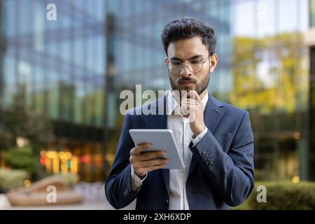 Young businessman in suit using digital tablet while standing outside modern office building. He looks thoughtful and focused on work in urban setting with glass reflections and greenery. Stock Photo
