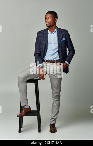 Handsome African American man in checkered blazer sitting on stool in formal attire against grey background. Stock Photo