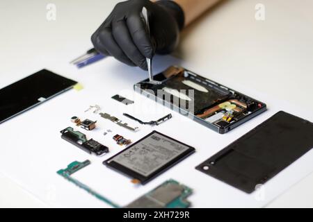 Professional repair of a disassembled smartphone, master with tweezers and gloves working on broken parts. Disassembled smartphone with parts laid out Stock Photo