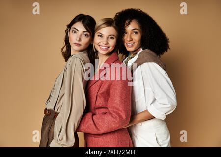 Three young, beautiful women representing different cultures stand together in trendy outfits on a beige background. Stock Photo
