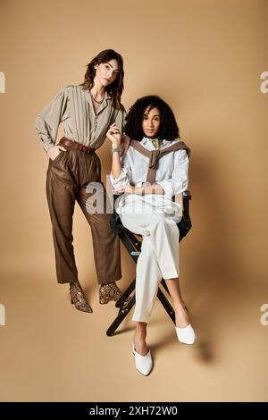 Two young, beautiful multicultural women in stylish attire, sitting on a chair, striking a pose for a picture. Stock Photo