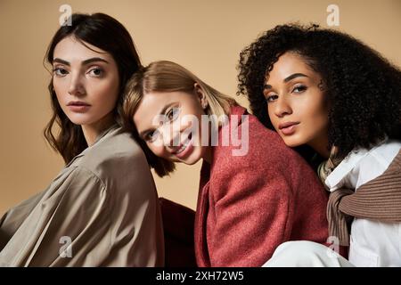 Three young, beautiful, multicultural women in stylish attire embracing their diversity while sitting together on a beige background. Stock Photo