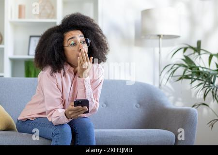 Young woman feeling bored at home sitting on couch holding smartphone. Casual indoor setting highlighting boredom and relaxation Stock Photo