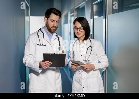 Medical professionals discussing patient records in hospital corridor. Man and woman in white coats with stethoscope and clipboard having conversation. Medical consultation and teamwork. Stock Photo